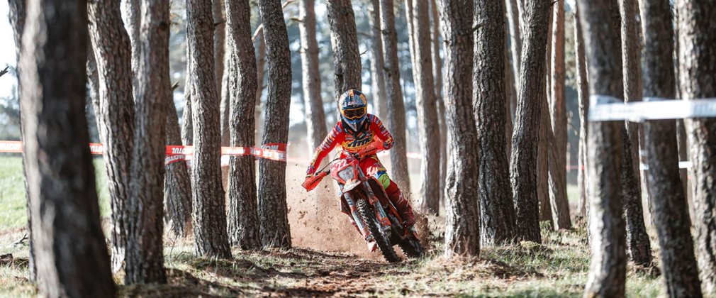 A person in full protective gear rides a motocross bike on a dirt track through a forest