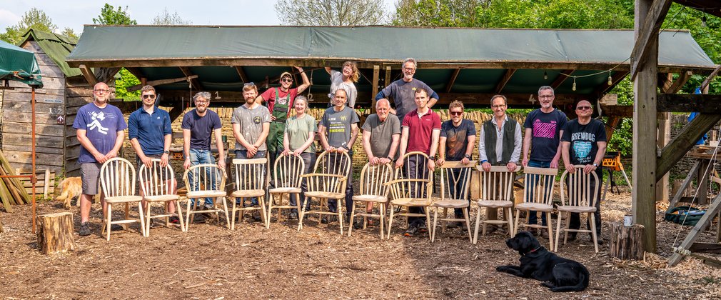 A group of people stand behind the wooden chairs they have made on a woodworking course.