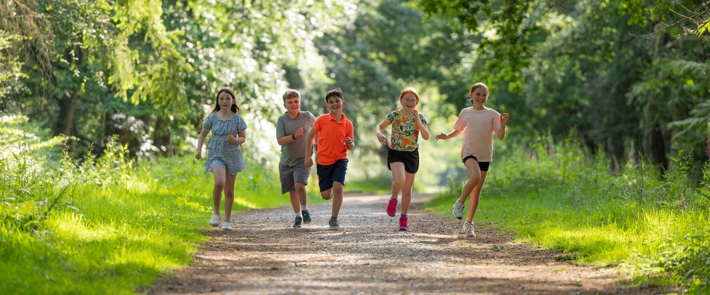 A summer image of five children running on a forest trail surrounded by trees in with dappled sunlight.
