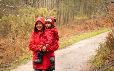 A woman in a red coat with hood up carries a child, wearing a red rain suit, along a forest path