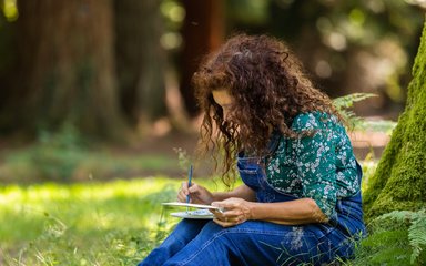 Woman sat at the base of a mossy tree drawing in a notebook