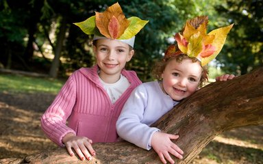 Two young girls, one in pink top and the other in white, wearing leaf crowns, standing at a tree branch and smiling.