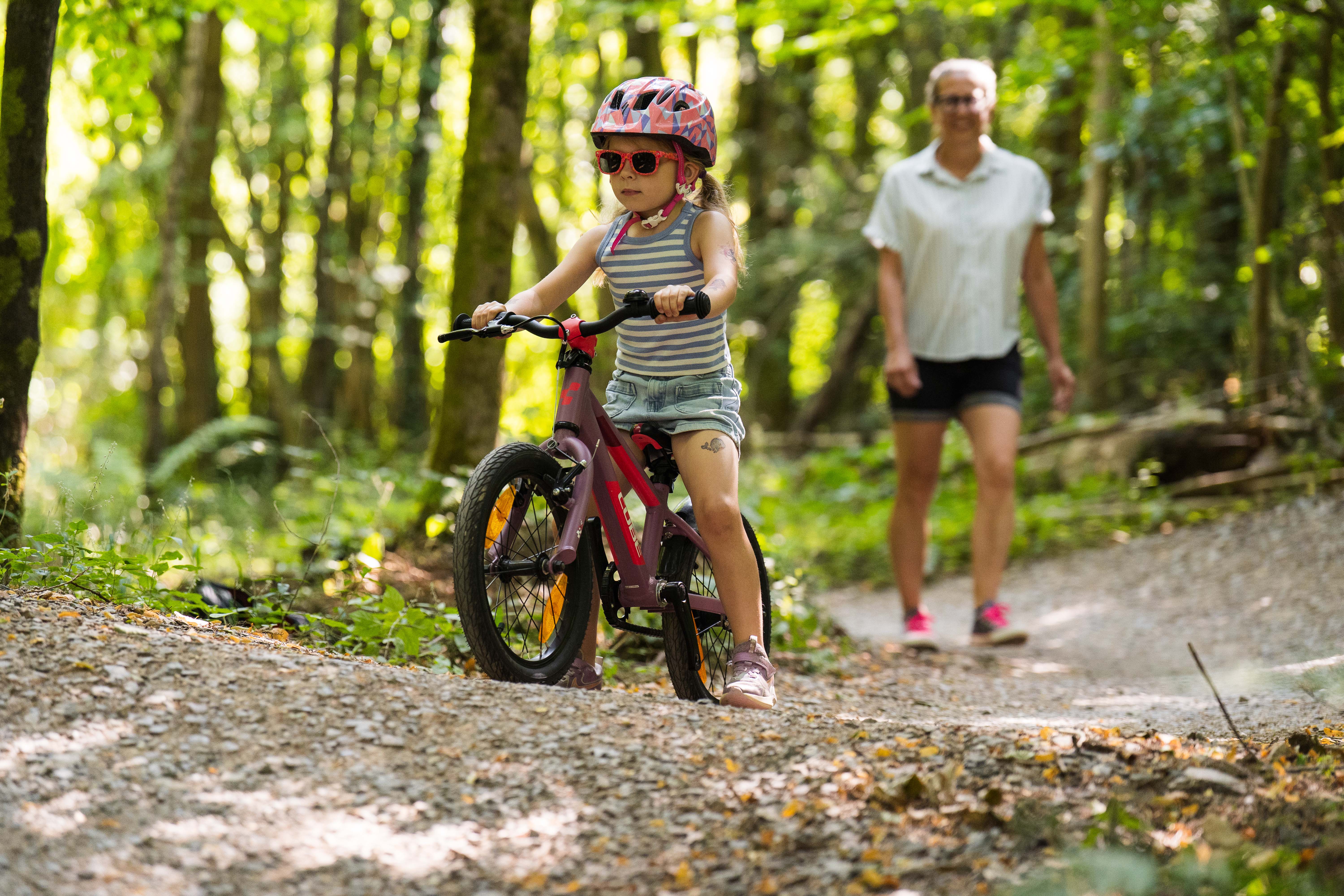 A young child rides her bike on a forest cycle trail while her parent follows behind