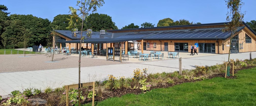 a single story long building stands in the background with a grassy area in the foreground. Chairs are dotted around outside in front of tables. A beautiful blue sky fills above the building