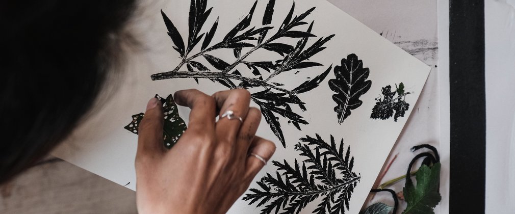 A piece of white paper has black leaf printings with a hand pressing down to ink the last leaf in place.