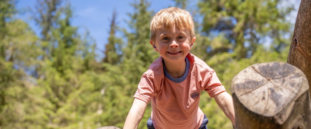 A boy on a wooden climbing frame smiling at the camera with trees in the background