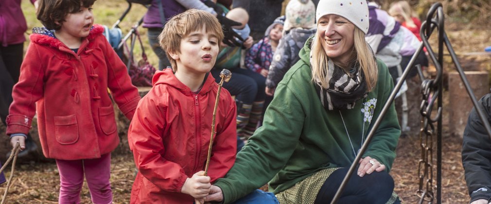 A lady and two children are crouching by a campfire toasting marshmallows