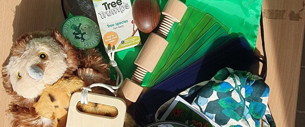 Contents of a sensory bag laid out on a green Forestry England bag. Items include a fluffy owl toy, wooden sensory toys, tree trumps, a magnifying glass, headphones. 