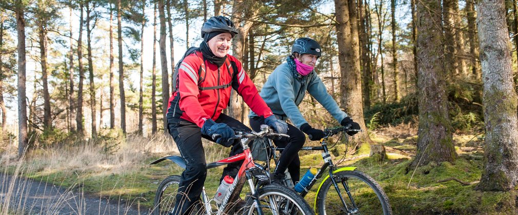 A man and a woman cycling through conifers