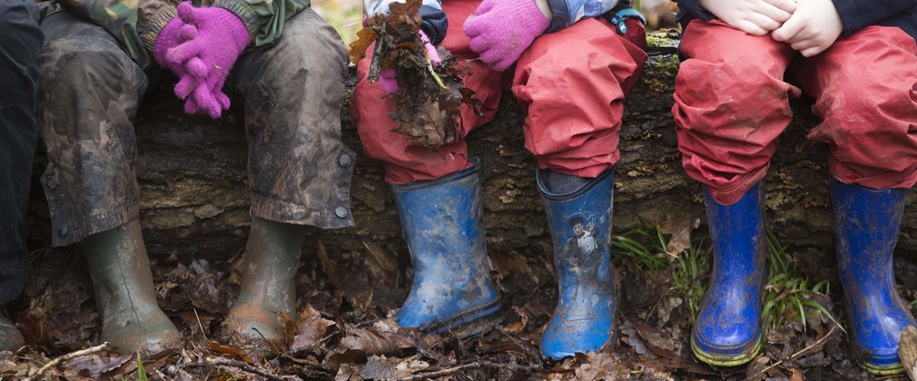 A close up of three children sitting on a log, showing legs in waterproof trousers and muddy wellington boots.