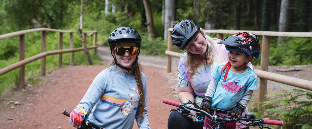 A woman and two young girls on mountain bikes smiling at the camera, ready to head out on a cycling trail.