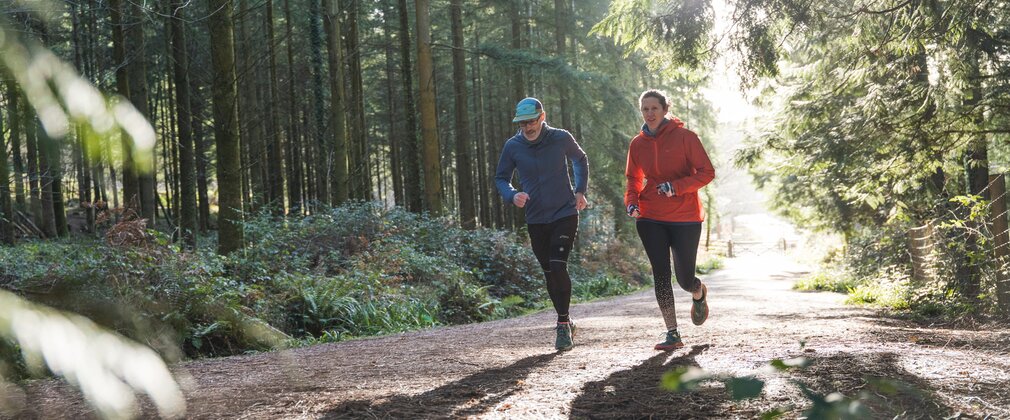 Two people running along a wide forest trail, with sunlight streaming through tall trees and casting long shadows across the path.