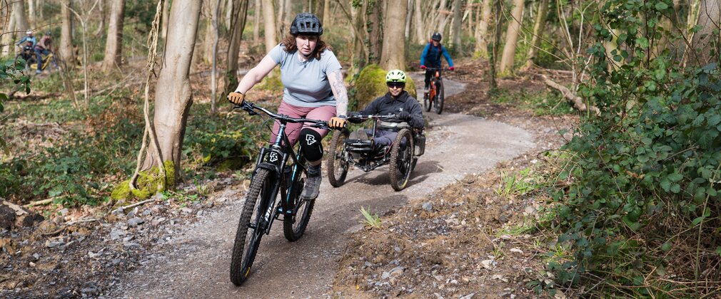 Three riders on different types of bike riding an accessible blue grade forest cycle trail