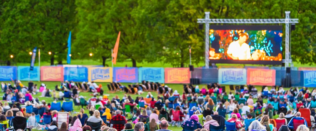 A large screen sits in front of a landscape of trees with Adventure cinema projected in the middle. Lots of people sit in deck chairs in front of the screen.
