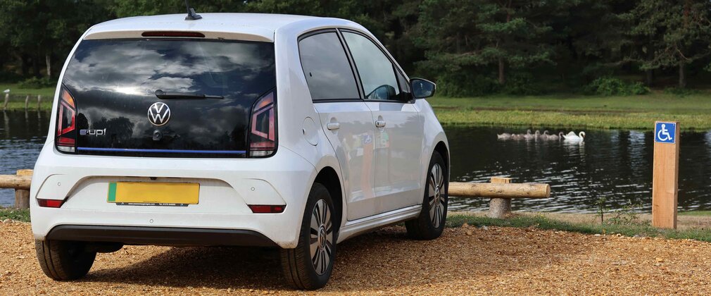 A white car parked in an accessible bay in a gravel car park beside a pond in the New Forest