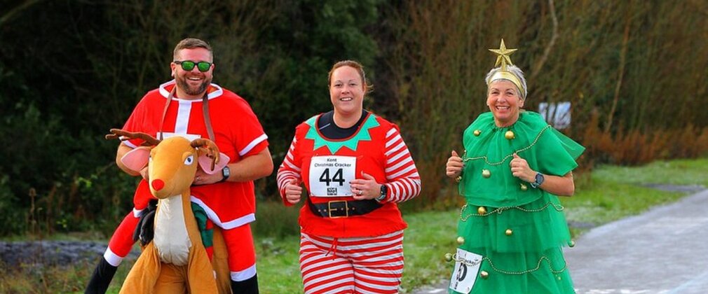 Three people running in a forest dressed as a reindeer, elf and Christmas tree