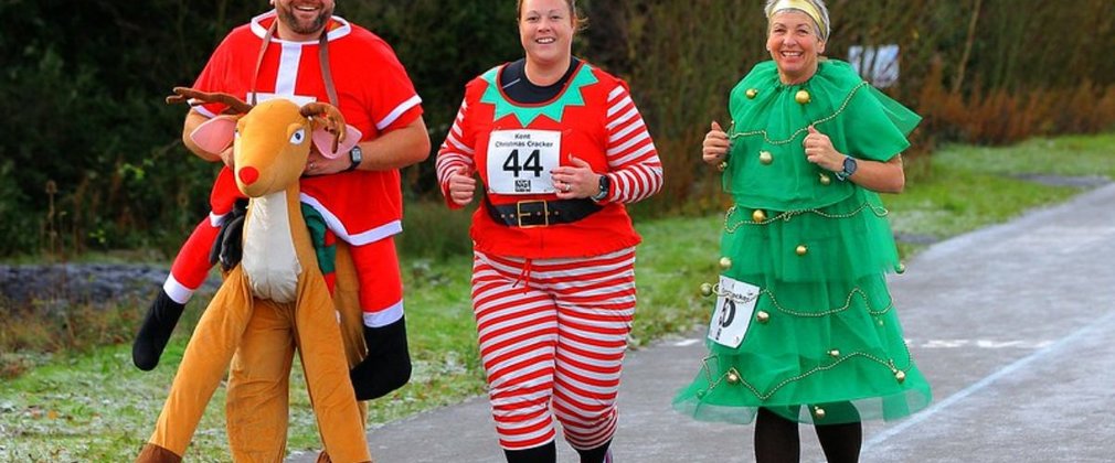 Three people running in a forest dressed as a reindeer, elf and Christmas tree