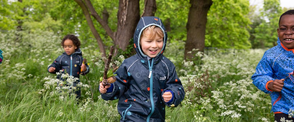 Three children in field with forest backdrop. One child is holding a stick.