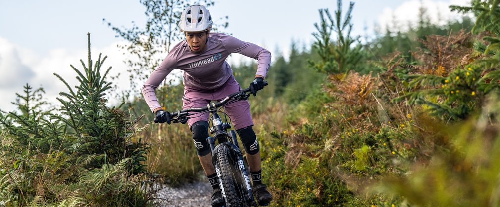 A mountain bike rider on a forest cycle trail. The rider is wearing pink clothes and a white helmet, cycling through young conifer trees.