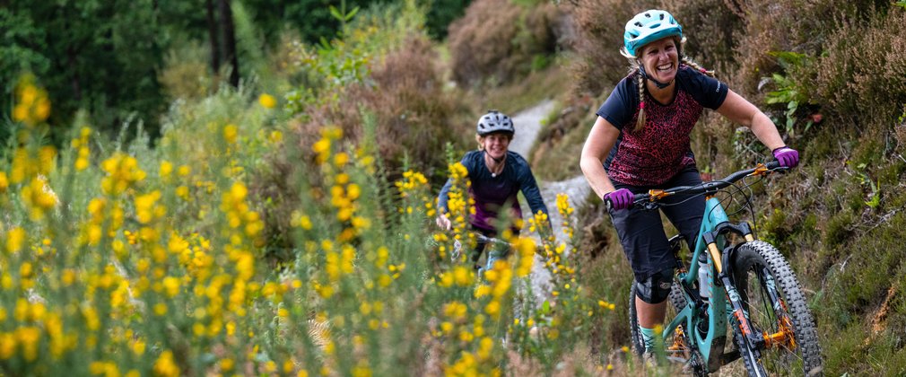 Two women on mountain bikes on a gravel track surrounded by summer foliage