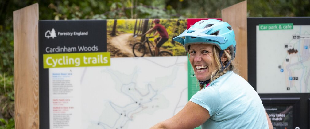 A female cyclist sitting on her bike and smiling over her shoulder with a map board in the background