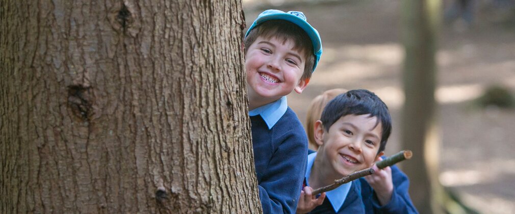 Two young boys in school uniform smile as their peer around a large tree trunk.
