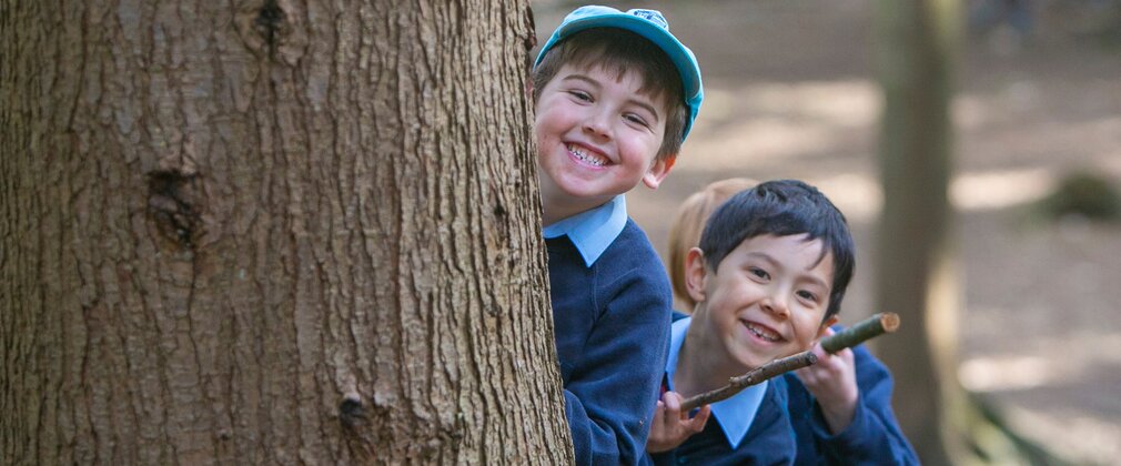 Two young boys in school uniform smile as their peer around a large tree trunk.