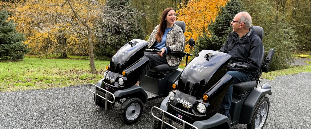A man and a woman driving two all terrain mobility scooters through a forest