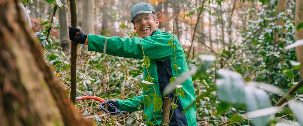 A man wearing a green Forestry England jacket in the woods, smiling at the camera and holding a handsaw and small branch
