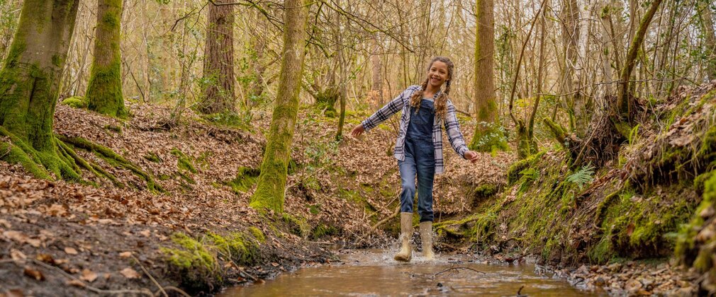 A girl wearing wellington boots splashing in a muddy puddle in the forest, with bare trees and leaves on the ground.
