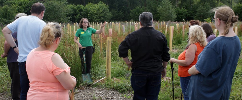 A woman dressed in green stands in front of a lot of tree planting tubes giving instructions to a group of people who have come to help. 