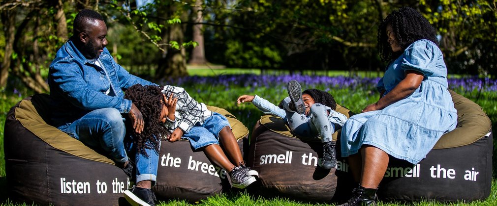 A family sit on large bean bags that have words saying listen to the birds, smell the air, feel the breeze. the children laugh and fall back on the beanbags, while the parents look relaxed and smile.