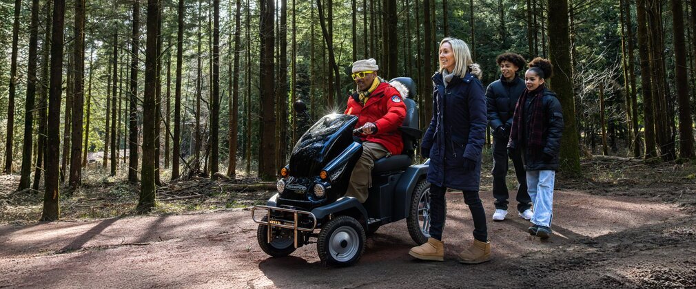 two adults and two children on a forest path, with one adult using an all-terrain mobility scooter.