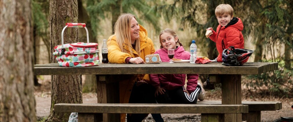 Mother and two children grinning while eating a picnic on a cluttered bench