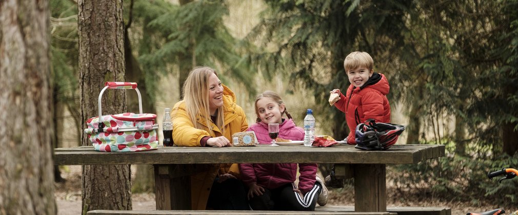 Mother and two children grinning while eating a picnic on a cluttered bench