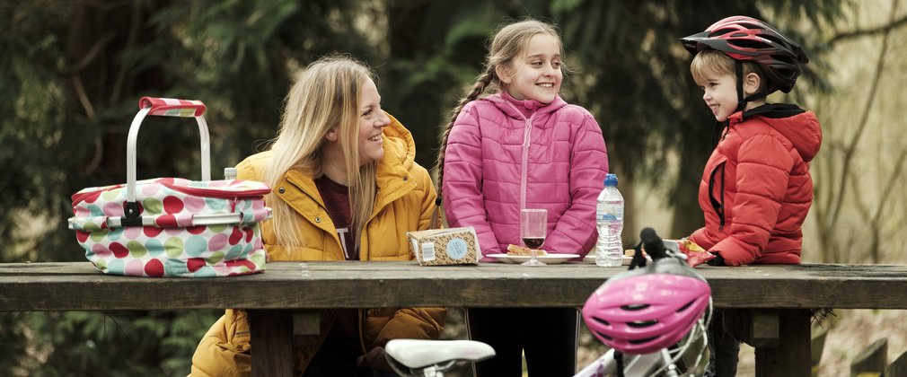 A young family sat on a wooden bench having a picnic in the forest