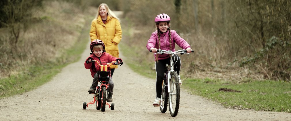 Woman behind two children, each of whom is riding a bike on a forest trail