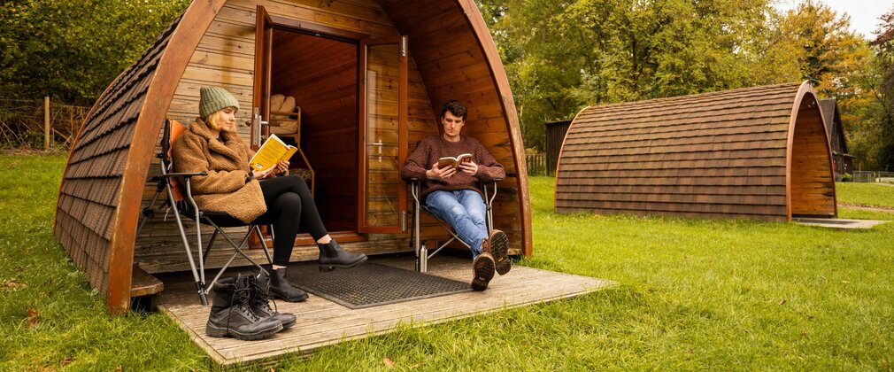 Wooden glamping pod with two people sitting outside reading books