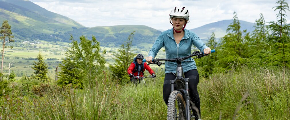 A woman in blue riding a mountain bike along a forest trail followed by a man in red