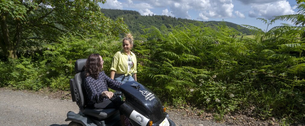 A youg woman on an off-road mobility scooter travels along a forest road whilst in conversation with another woman wearing yellow.