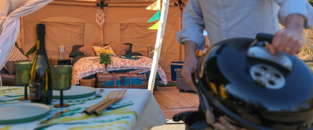 Man cooking sausages on a barbeque outside a furnished bell tent with bunting flags behind him