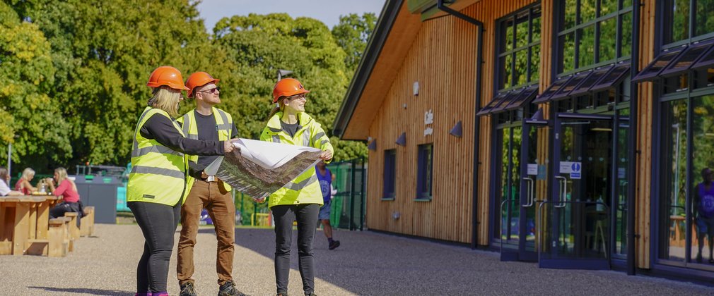 two women and a man in high vis clothing and hard hats looking at plans in front of a timber clad building