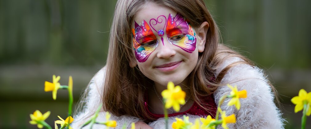 A girl with her face painted with a butterfly looks contently at some daffodils