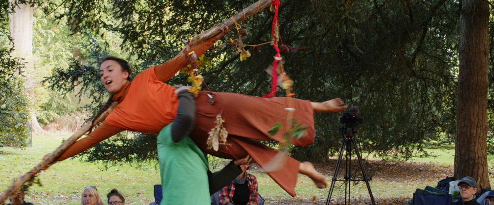 A woman dressed in bright orange is being lifted by a person in green, over their shoulder. She looks like she is flying through the air with a long stick attached to her arms.