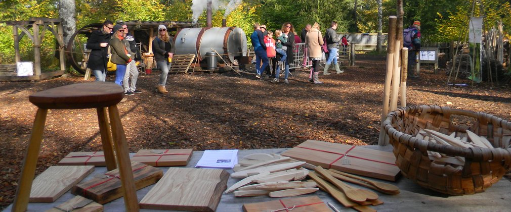In the forefront you see a table with wooden produce including a stool and tent pegs. In the background people are looking around at the coppice area.
