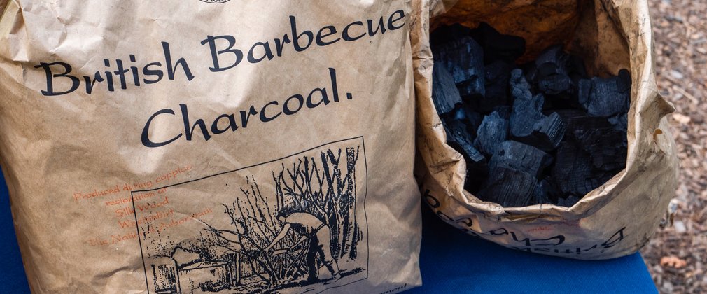 2 large bags of charcoal sit on a table, one displays the name 'British Barbecue Charcoal', and the other sits open to peek inside to see black pieces of charcoal