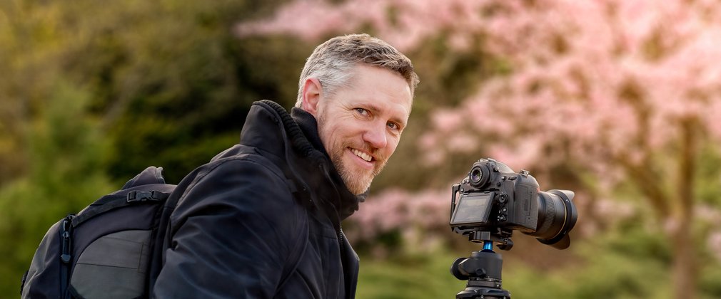 A man smiling at the camera with a large camera on a tripod on a spring day 
