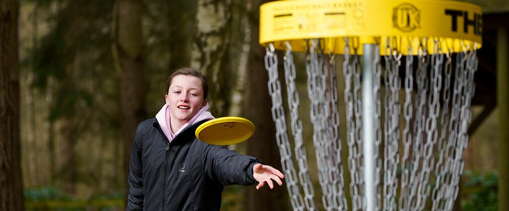 A young person throwing a yellow flying disc towards a disc golf net.