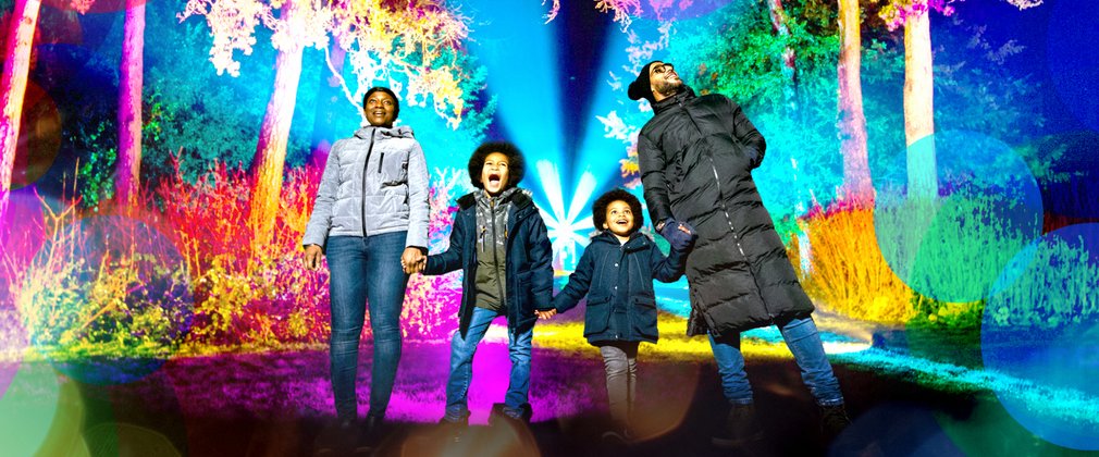 A family hold hands in a line looking in awe at brightly coloured lights of enchanted Chrismtas surrounding them.