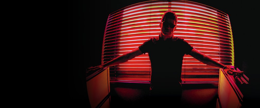 Noel Gallagher stands confidently in front of illuminated blinds 